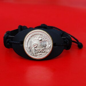 May include: A black leather bracelet with a silver coin featuring a woman looking at a rocket ship and the words "United States of America".