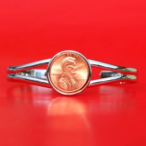 May include: A silver bracelet with a double band design features a 1983 US penny coin with the inscription "In God We Trust" and "Liberty". The coin is centered on the bracelet and is surrounded by a silver band.
