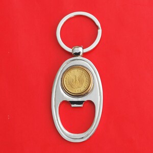 Puede incluir: Abrebotellas de llavero de plata con una moneda de oro que presenta un diseño de pájaro y el texto "ΔΡΑΧΜΗ 1873".