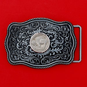 May include: A silver-toned belt buckle with an ornate design featuring a five-cent coin with the image of a buffalo. The coin reads "United States of America" and "Five Cents".