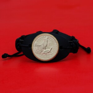 May include: A black leather bracelet featuring a gold-colored coin with an image of a wolf, turtle, and text that reads "TREATY WITH THE DELAWARES 1778". The bracelet has a braided cord closure and is set against a red background.
