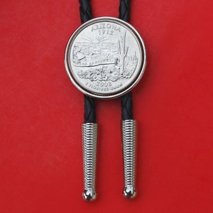 May include: A silver bolo tie featuring a 2008 Arizona state quarter with the text "Arizona 1912" and "Grand Canyon State" on the front. The tie has black braided cords with silver tips.