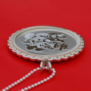 May include: A silver pendant necklace featuring a circular charm with an engraved design of Mickey Mouse and Minnie Mouse. The charm has a scalloped edge and is attached to a silver chain.