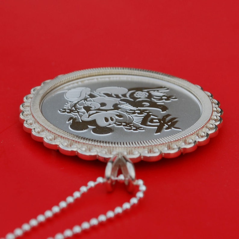 May include: A silver pendant necklace featuring a circular charm with an engraved design of Mickey Mouse and Minnie Mouse. The charm has a scalloped edge and is attached to a silver chain.
