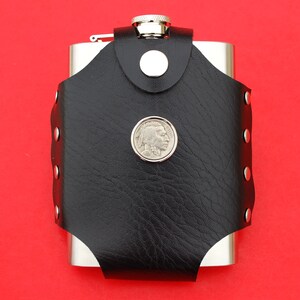 May include: A silver flask with a black leather cover and a silver coin with a profile of a Native American on it. The flask has a hinged lid and a small loop for carrying.