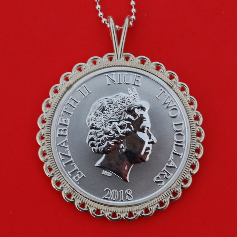 May include: Silver coin pendant with a portrait of Queen Elizabeth II and the text "NIUE TWO DOLLARS 2018". The coin is surrounded by a decorative gold-colored border.