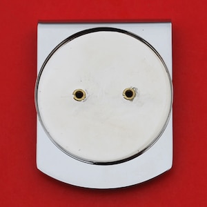 May include: A polished silver money clip with a circular, recessed area. The circle has two small, gold-colored rivets. The clip is rectangular with rounded edges and a shiny finish. The background is a solid red.