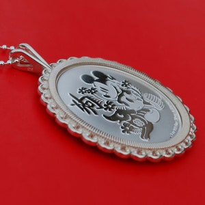 May include: Silver pendant featuring a Mickey Mouse and Minnie Mouse design with Chinese characters. The pendant is on a red background.