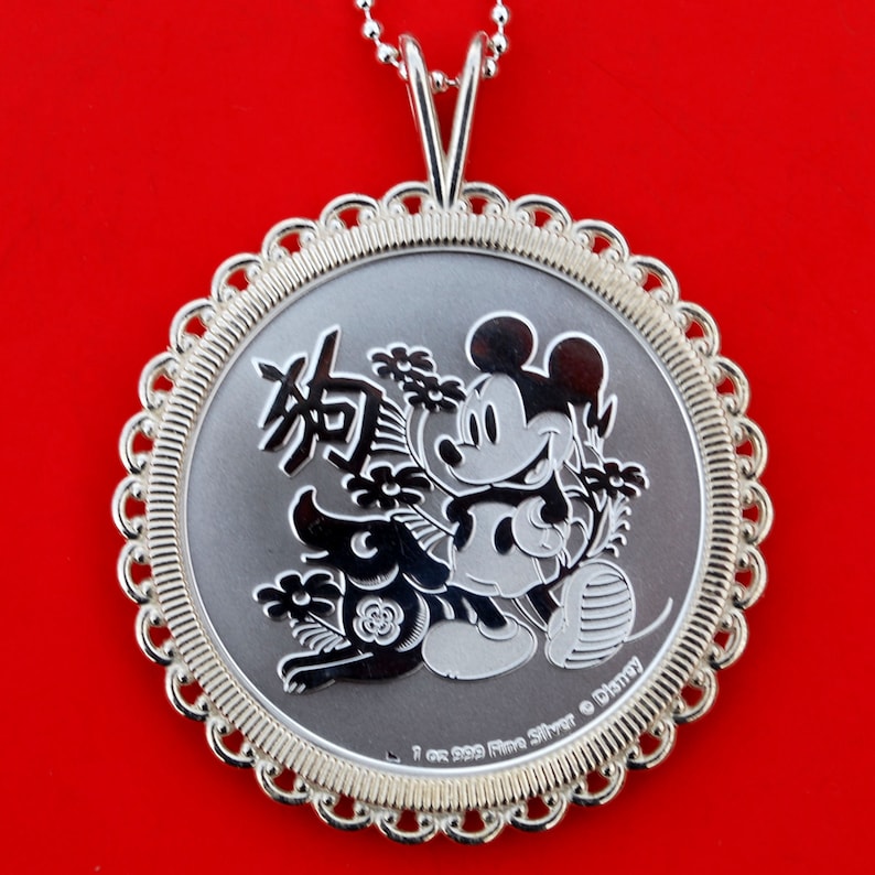 May include: A silver pendant necklace featuring Mickey Mouse and the Chinese character for 'dog' in a circular design. The pendant is surrounded by a decorative border and has the inscription '1 oz 999 Fine Silver &copy; Disney'.