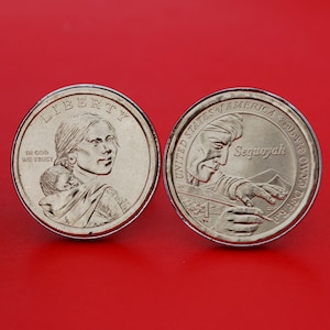 May include: Two silver US dollar coins. The obverse side features a portrait of a woman with a child on her back. The inscription reads "Liberty" and "In God We Trust". The reverse side features a portrait of Sequoyah, a Cherokee silversmith, and the inscription "United States of America" and "One Dollar".
