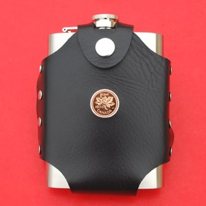 Puede incluir: Una petaca de acero inoxidable con una cubierta de cuero negro y una moneda canadiense de 1 centavo de 1970 unida al frente.