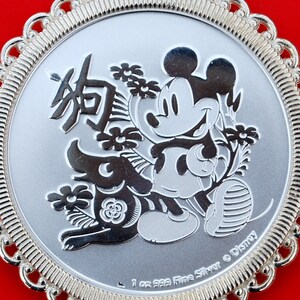 May include: A silver coin with a Mickey Mouse design celebrating the Chinese Year of the Dog. The coin features Mickey Mouse standing next to a dog, with flowers and the Chinese character for 'dog' in the background. The coin is inscribed with "1 oz 999 Fine Silver &copy; Disney".