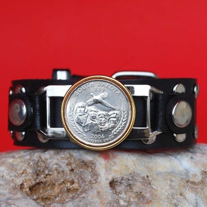 May include: A black leather bracelet with silver studs and a silver buckle. The buckle features a 2006 South Dakota quarter with a gold rim. The coin depicts a pheasant and the Mount Rushmore National Memorial.