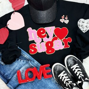May include: Black sweatshirt with the text "hey sugar" in red and pink glitter letters. A red heart with glitter is next to the text. The sweatshirt is paired with blue jeans and black sneakers.