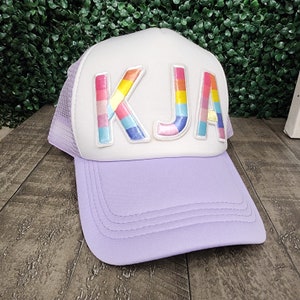 May include: A white and purple trucker hat with a rainbow-colored felt patch that says "KJA" on the front.