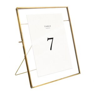 May include: Gold metal picture frame with a clear glass front. The frame is standing upright on a white background. The frame holds a white card with the text "TABLE 7" printed on it.