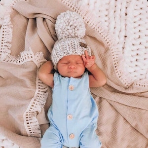 May include: A baby wearing a light blue onesie with buttons and a gray knitted hat with a pom-pom on top. The baby is lying on a white and beige blanket.