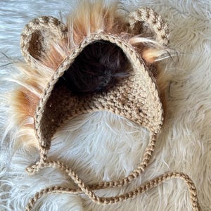 May include: A tan crochet lion hat with faux fur mane and ears. The hat has a rounded crown and ear flaps with braided ties. The faux fur is a mix of brown, tan, and orange. The hat is designed for babies and toddlers.