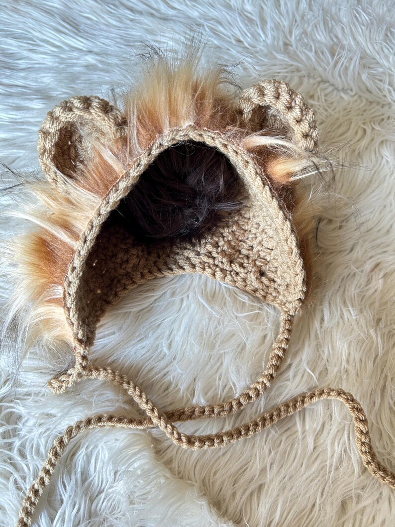 May include: A tan crochet lion hat with faux fur mane and ears. The hat has a rounded crown and ear flaps with braided ties. The faux fur is a mix of brown, tan, and orange. The hat is designed for babies and toddlers.