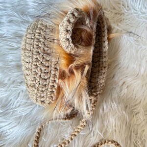 May include: A tan crocheted baby hat with faux fur trim. The hat features a lion's mane design with a braided chin strap. The fur is a mix of light brown and tan, adding a playful touch to the handmade accessory.