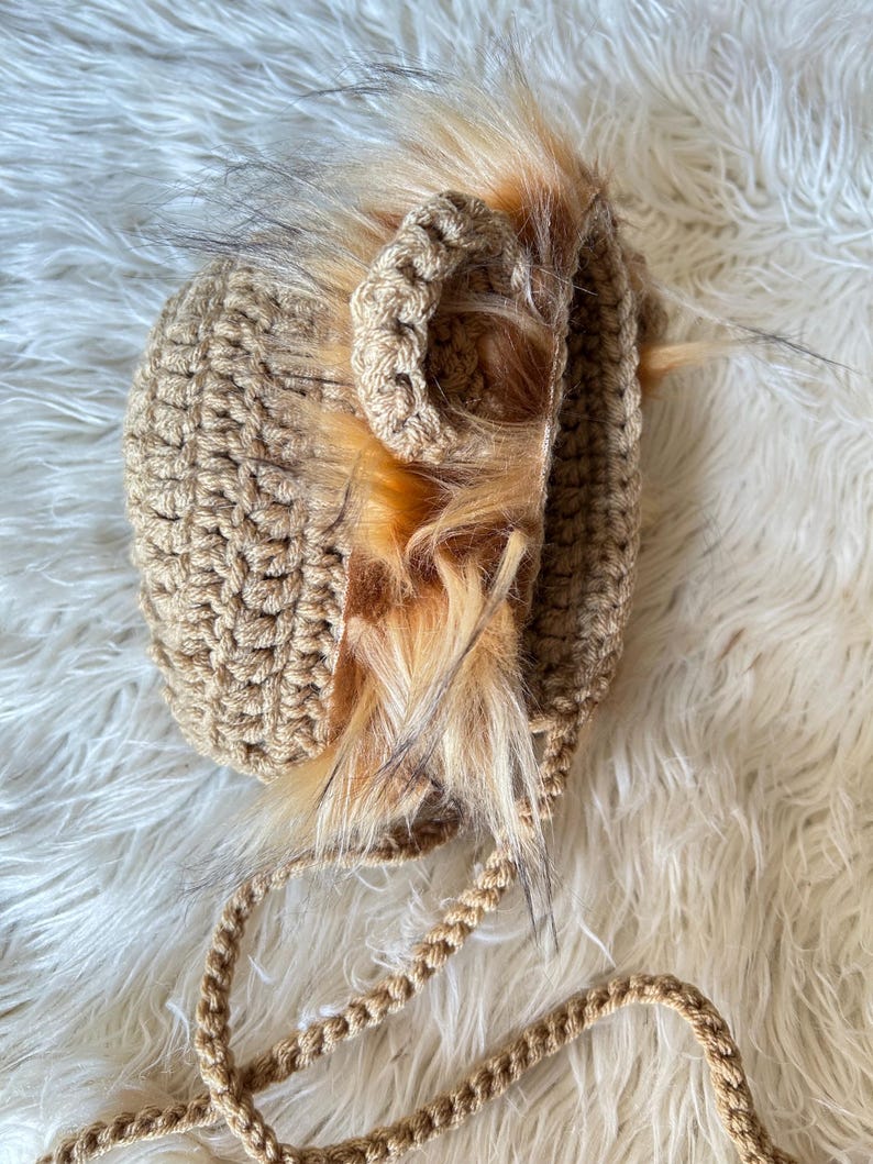 May include: A tan crocheted baby hat with faux fur trim. The hat features a lion's mane design with a braided chin strap. The fur is a mix of light brown and tan, adding a playful touch to the handmade accessory.