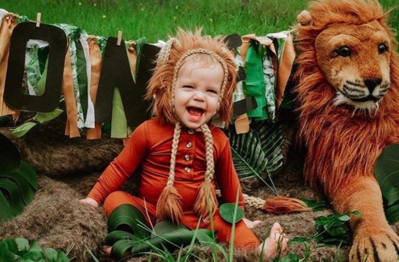 May include: A smiling child in an orange lion costume with a faux fur mane and braids. The child is sitting in a jungle-themed setting with a large stuffed lion and a banner that says "ONE".