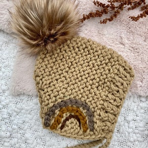 May include: A tan crocheted baby hat with a brown faux fur pom pom and a rainbow embroidery detail.