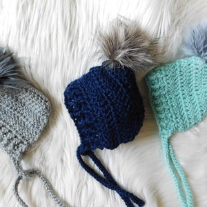 May include: Three crocheted baby hats with faux fur pom poms. The hats are in grey, blue, and green.