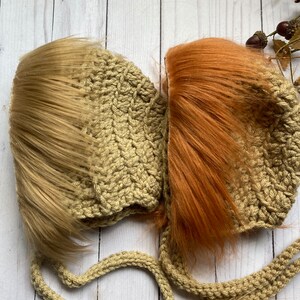 May include: Two handmade crochet baby hats, one with light brown faux fur and the other with orange faux fur. The hats are a light tan color and have long ties. The hats are designed to look like a lion's mane.
