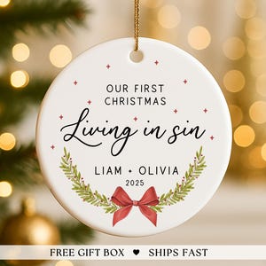 May include: A white ceramic Christmas ornament with gold string. The ornament features the text "OUR FIRST CHRISTMAS Living in sin" with the names "LIAM + OLIVIA" and the year "2025". A red bow and green wreath complete the design.