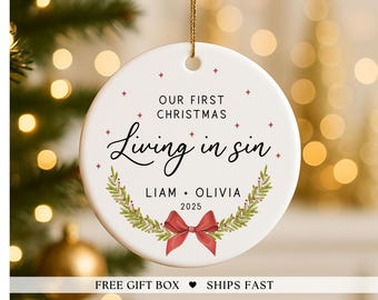Könnte beinhalten: Weißer Keramik-Weihnachtsschmuck mit goldener Schnur. Der Schmuck zeigt den Text "OUR FIRST CHRISTMAS Living in sin" mit den Namen "LIAM + OLIVIA" und dem Jahr "2025". Ein roter Bogen und ein grüner Kranz ergänzen das Design.