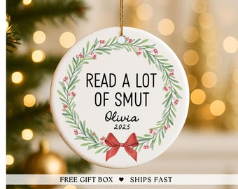 May include: A white ceramic Christmas ornament with the text "READ A LOT OF SMUT" in black, surrounded by a green and red wreath. The name "Olivia" and the year "2025" are also included. A red bow is at the bottom.