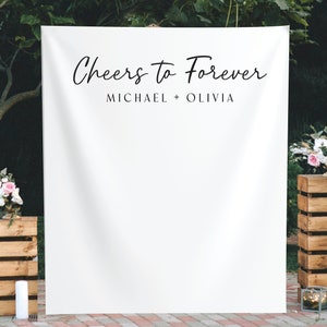K&ouml;nnte beinhalten: Wei&szlig;er Hintergrund mit dem Text "Cheers to Forever" und den Namen "Michael + Olivia" in schwarzer Schrift. Der Hintergrund ist im Freien mit Holzkisten und Blumen aufgebaut.