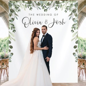 May include: A wedding photo backdrop with a white background and green foliage border. The text "THE WEDDING OF Olivia & Josh" is printed in black at the top of the backdrop.