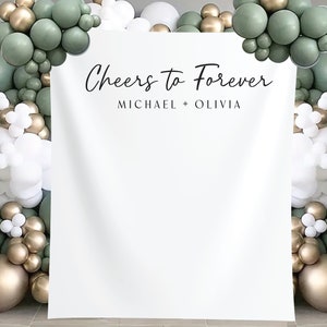 K&ouml;nnte beinhalten: Wei&szlig;er Hintergrund mit dem Text "Cheers to Forever" und "Michael + Olivia" f&uuml;r eine Hochzeits- oder Verlobungs-Fotobox. Der Hintergrund ist mit einer Girlande aus gr&uuml;nen, goldenen und wei&szlig;en Ballons dekoriert.