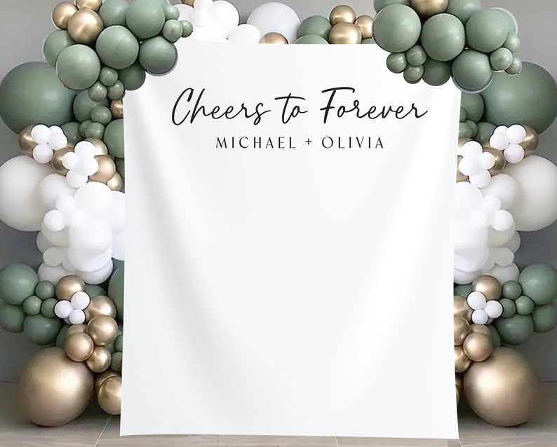 K&ouml;nnte beinhalten: Wei&szlig;er Hintergrund mit dem Text "Cheers to Forever" und "Michael + Olivia" f&uuml;r eine Hochzeits- oder Verlobungs-Fotobox. Der Hintergrund ist mit einer Girlande aus gr&uuml;nen, goldenen und wei&szlig;en Ballons dekoriert.
