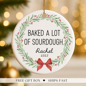 May include: A white ceramic Christmas ornament with the text "BAKED A LOT OF SOURDOUGH" in black, surrounded by a green and red wreath. The name "Rachel" and the year "2025" are below the text, with a red bow at the bottom.