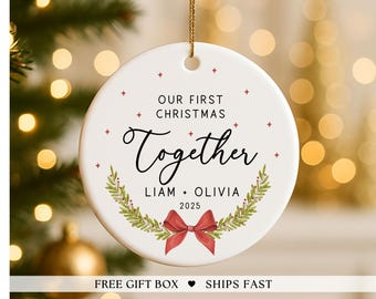May include: A white ceramic Christmas ornament with the text "OUR FIRST CHRISTMAS TOGETHER" in black script. The names "LIAM + OLIVIA" and the year "2025" are also included. The ornament features a red bow and a green wreath design.