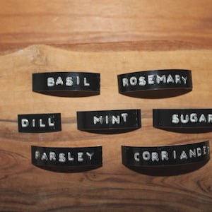 Op de afbeelding: Zwarte, gebogen kruidenlabels met witte letters voor basilicum, rozemarijn, dille, munt, suiker, peterselie en koriander. De labels zijn gerangschikt op een houten snijplank.