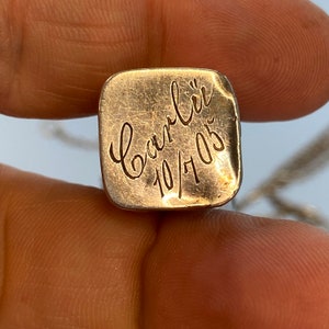 May include: A square, gold-colored pendant with rounded corners. The pendant has the name "Carli" and the date "10/7/05" engraved in a cursive script. The pendant is held between two fingers.
