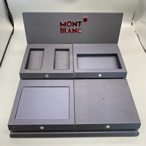 May include: A grey Montblanc presentation box with compartments. The box has a hinged lid with the Montblanc logo in red and white. The interior has several compartments for storing items. The box is made of a sturdy material.