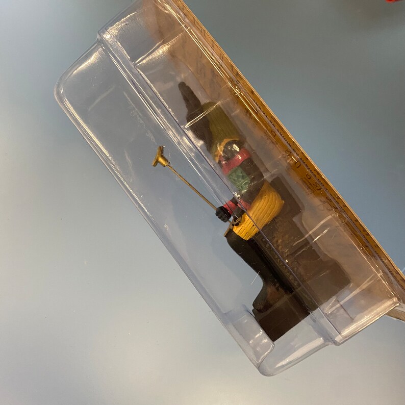Peut inclure: Une petite figurine color&eacute;e repr&eacute;sentant une personne assise sur un banc, tenant un b&acirc;ton. La figurine est en bois et peinte dans des tons de vert, rouge, jaune et noir. La figurine est emball&eacute;e dans une coque en plastique transparente.