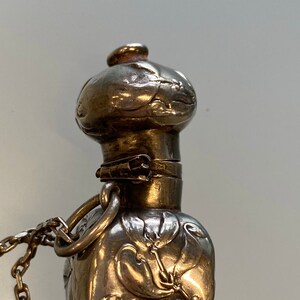 May include: Close-up of a silver-toned perfume bottle with floral detailing. The bottle has a bulbous shape with a decorative top and a small chain attached. The metal has a tarnished appearance, suggesting age.