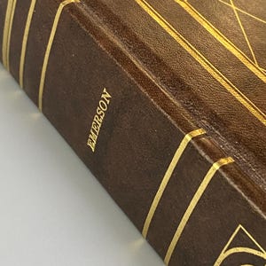 May include: Close-up of a vintage book with a brown leather cover and gold accents. The word "EMERSON" is embossed in gold on the spine. The book has gold-lined edges and decorative gold patterns.