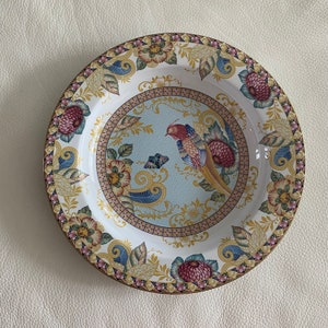 Könnte beinhalten: Ein weißer Keramikteller mit einem farbenfrohen Blumen- und Vogeldesign. Der Teller zeigt einen blauen und gelben Vogel mit roter Brust, umgeben von Blumen und Blättern in den Farben Rosa, Rot, Gelb und Grün. Der Teller hat einen goldenen Rand.