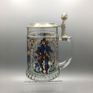 Könnte beinhalten: Ein Bierkrug aus klarem Glas mit silbernem Deckel und Griff. Der Krug zeigt eine farbenfrohe Illustration eines Mannes in historischer Kleidung, umgeben von floralen und architektonischen Elementen. Das Design erinnert an Buntglas.