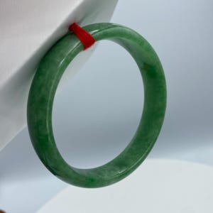 May include: A green jade bangle bracelet with a smooth, polished finish. The bangle is thick and has a rounded shape.