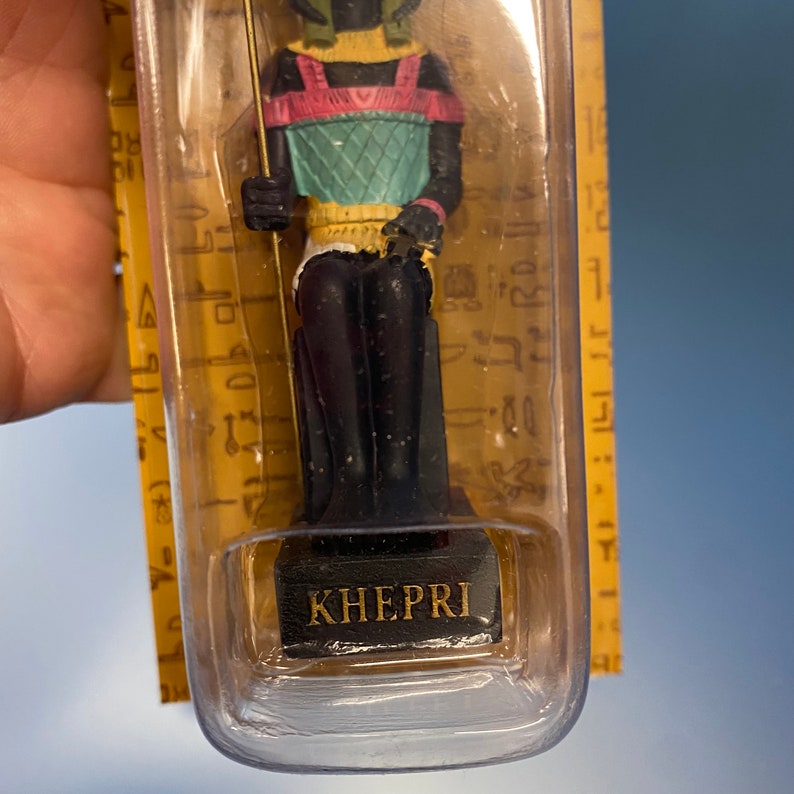 Peut inclure: Une petite figurine en plastique du dieu &eacute;gyptien Kh&eacute;pri, repr&eacute;sent&eacute; comme un scarab&eacute;e avec une t&ecirc;te humaine. La figurine porte une tunique color&eacute;e et tient un b&acirc;ton. La figurine est pr&eacute;sent&eacute;e dans un bo&icirc;tier en plastique transparent avec une base noire sur laquelle est inscrit "KHEPRI".