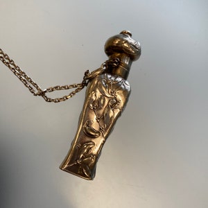 May include: A silver-toned perfume bottle with floral relief detailing and a decorative cap. The bottle is suspended from a delicate chain. The design features stylized flowers and leaves, creating an elegant, vintage aesthetic.