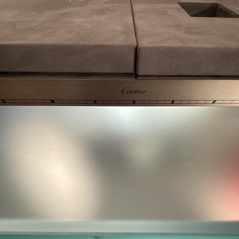 May include: A silver metal box with a brown suede top and the word 'Cartier' engraved on the front.